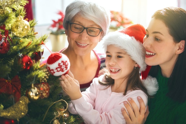 Frohe Weihnachten und schöne Feiertage! Oma, Mama und Tochter schmücken den Baum drinnen. Der Morgen vor Weihnachten. Porträt liebevolle Familie hautnah.