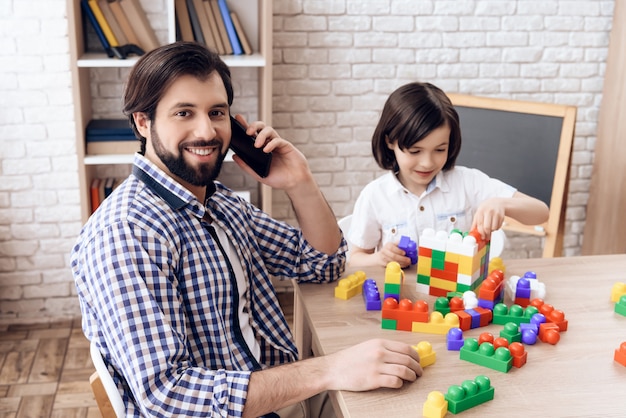 Froher Vater spricht am Telefon, während Sohn mit Spielzeug spielt.