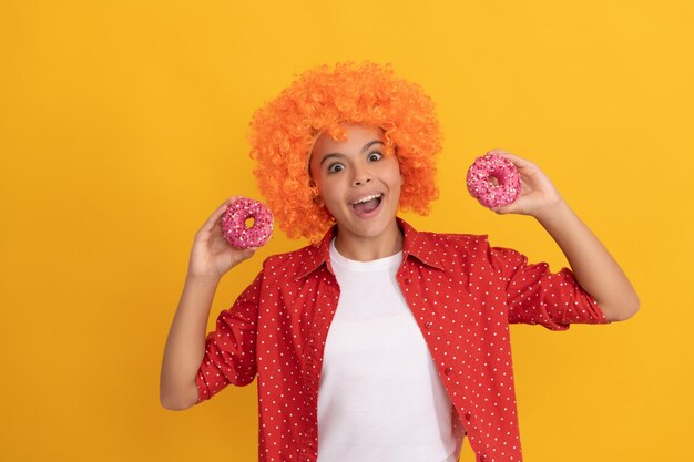 frohes jugendlich Mädchen in schicker orangefarbener Haarperücke hält süß gebackenen Donut-Dessert, Zahnpflege.