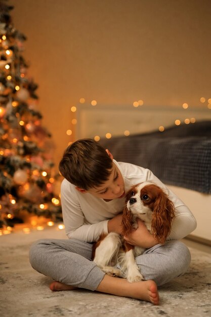 Frohes neues Jahr ein Junge in einem leichten hausgemachten Pyjama umarmt sein Haustier Cavalier King Charles Spaniel zu Hause im Schlafzimmer in der Nähe des Weihnachtsbaums