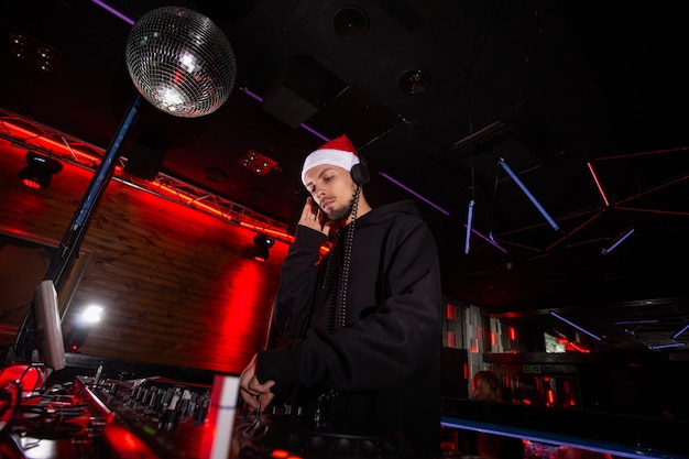 Frohes neues Jahr und frohe Weihnachten Charismatischer Discjockey in rotem Weihnachtsmann-Hut, Kopfhörer und Kapuze spielt Musik an DJ-Plattenspielern. Partykonzept für das neue Jahr.