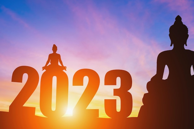 Frohes neues Jahr Zahlen 2023 Silhouette Buddha-Statue und Silhouette Frau praktizieren Yoga am frühen Morgen Sonnenaufgang über dem Horizont HintergrundFrohes neues Jahr Konzeptlebendige Dämmerung dramatischer Sonnenuntergang