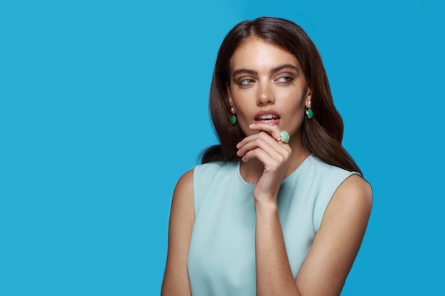 Frontalporträt einer jungen Frau in hellblauem Kleid mit eleganten Accessoires. Schmuck-Ohrringe, Schmuck-Ring, auf blauem Hintergrund.