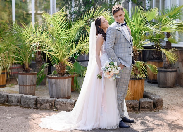 Frontansicht des eleganten Brautmädchens in Hochzeitskleid und Schleier, das einen Blumenstrauß hält, der hinter einem hübschen Bräutigam steht, der in einem botanischen Garten posiert Hochzeitstag für ein Liebespaar