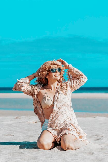Frontansicht einer schönen Frau mit offenen Armen, die am Strand sitzt. Blonde Frau mit lockigem Haar, die im Wind weht, entspannt sich, schaut und lächelt.