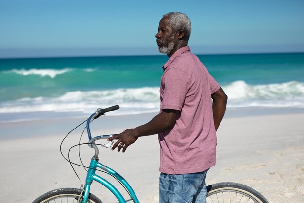 Frontansicht eines älteren afroamerikanischen Mannes, der am Strand mit blauem Himmel und Meer im Hintergrund steht, ein blaues Fahrrad hält und wegblickt