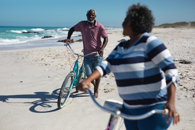 Frontansicht eines älteren afroamerikanischen Paares, das am Strand mit blauem Himmel und Meer im Hintergrund steht, ihre Fahrräder fährt und sich gegenseitig anschaut und lächelt