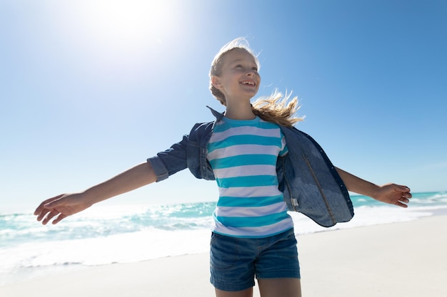 Frontansicht eines kaukasischen Mädchens auf einem sonnigen Strand, lächelnd mit ausgestreckten Armen, mit blauem Himmel und Meer im Hintergrund