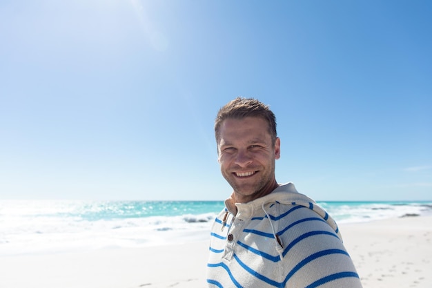 Frontansicht eines kaukasischen Mannes, der am Strand mit blauem Himmel und Meer im Hintergrund steht und zur Kamera lächelt