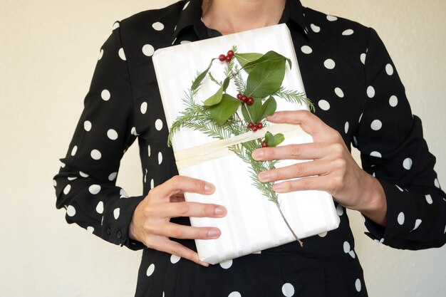Frontaufnahme einer nicht erkennbaren Frau, die ein Weihnachtsgeschenk mit Weihnachtspflanzendekoration hält, heller Hintergrund