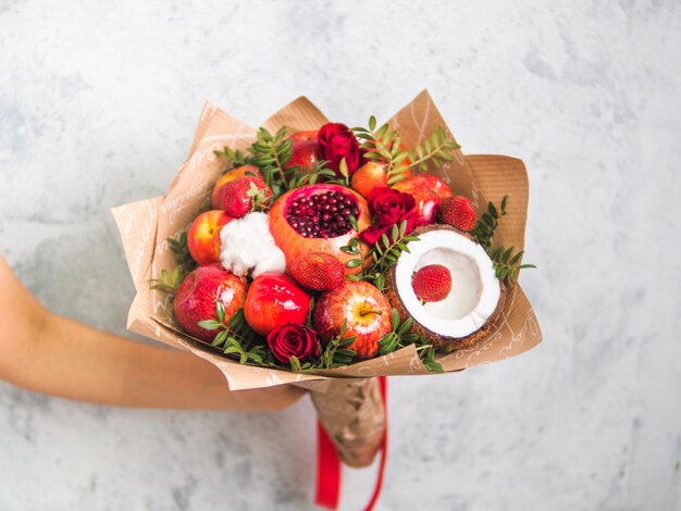 Frucht- und Beeren-Bukett Essen-Bukett in weiblicher Hand auf grauem Hintergrund Granatapfel Erdbeere Äpfel Kokosnuss und Rosen Blumen Eukalyptus Flaches DOF Kopierraum