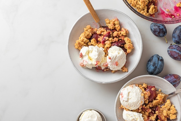 Fruchtstreusel. Pflaumenstreuselkuchen mit Vanilleeis in Tellern mit Löffeln auf weißem Marmortisch für leckeres Frühstück. Ansicht von oben