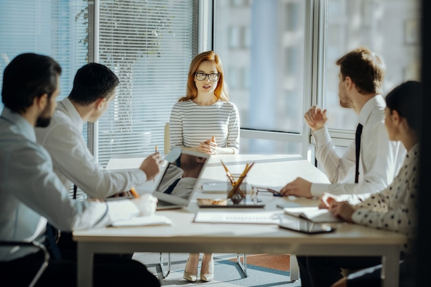 Fructífera colaboración. Encantadora jefa sentada a la cabecera de la mesa con sus empleados y desarrollando un plan de proyecto junto con ellos