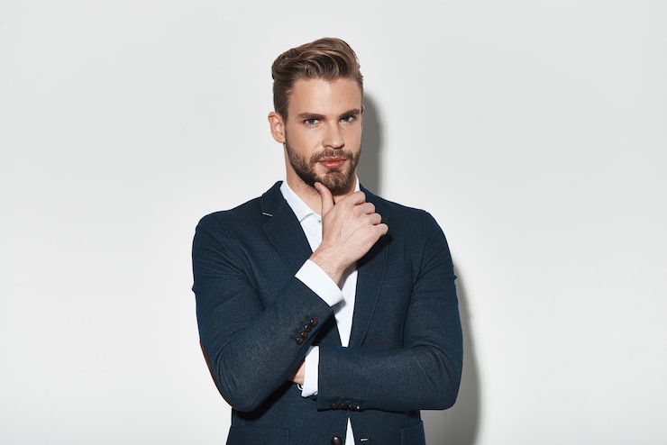 Foto früher sah es perfekt aus. hübscher junger mann im vollen anzug, der die hand am kinn hält und die kamera ansieht, während er vor grauem hintergrund steht
