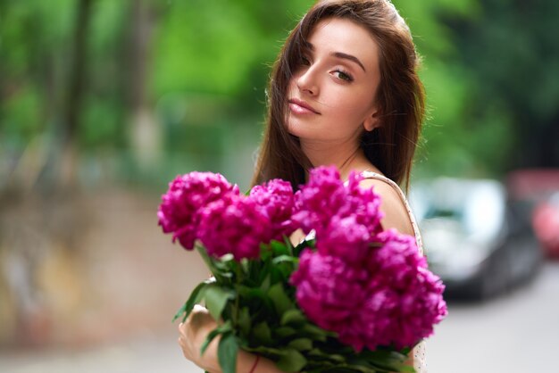 Frühlings- / Sommerstil. Schöne junge brünette Frau in einem schönen Frühlingskleid mit einem Blumenstrauß von Pionen. Schöne Frühlingsstraße. Mode Frühling Sommer Foto.