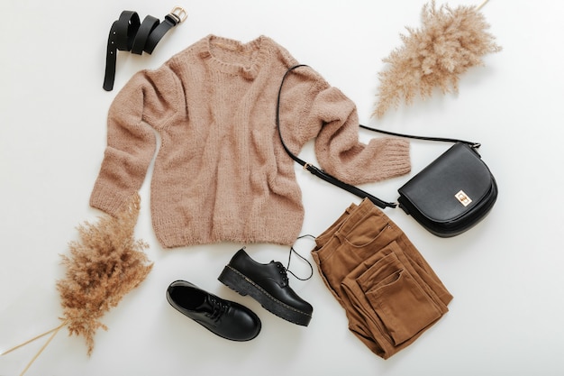 Frühlings-Winter-Herbst-Frauen-Outfit. Mode-Basic-Frauenkleidung in beige-brauner Farbe. Stoff und Schuhe lässiger Stil mit Strickpullover und Schilf-Pampaszweigen. Flache Kleidung auf Weiß legen.