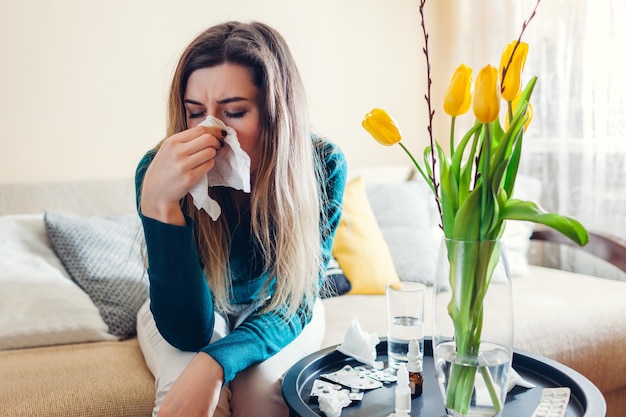 Frühlingsallergie Frau niest wegen Tulpenblumen, umgeben von Pillen und Tropfen zu Hause Saisonale Allergie