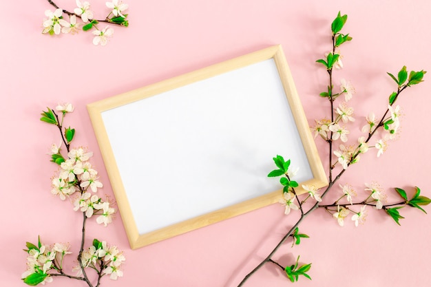 Frühlingsblumenzweige mit Kirschblüten um Fotorahmen. Weißer Rohling für inspirierenden oder motivierenden Text und Zitat auf weichem rosa Hintergrund. Modell, flache Draufsicht, Kopierraum.