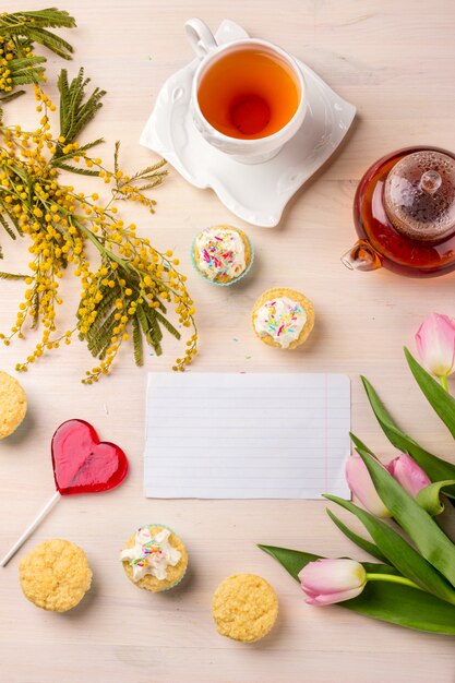 Frühlingsgrußkarte mit Tulpen, Mimose, Tee und kleinen Kuchen auf hölzernem Hintergrund.