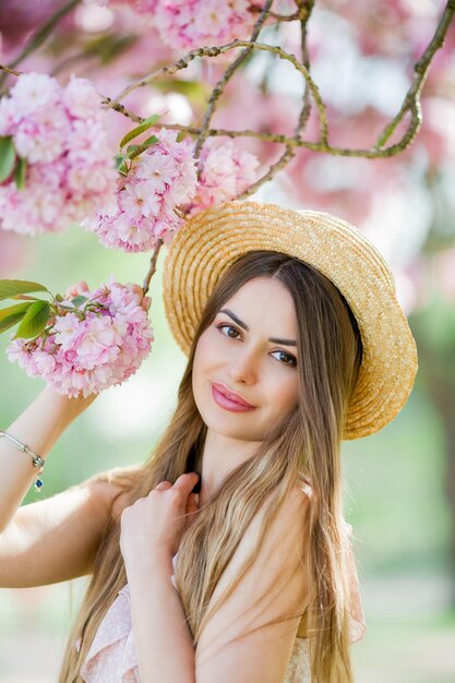 Frühlingsporträt einer jungen schönen Frau in rosa Blüten Junges schönes Modell mit langen Haaren in einem Weidenhut und einem leichten Kleid in der Nähe der Kirschblüten