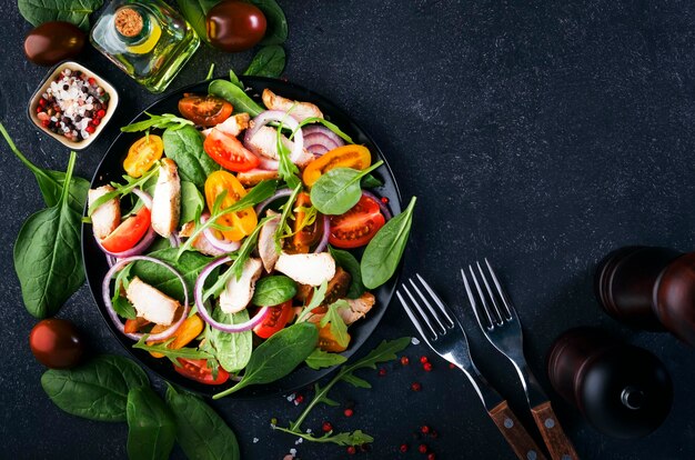 Frühlingssalat mit Spinat, gegrilltem Hähnchen, Kirschtomaten, Arugula, Maissalat und roten Zwiebeln, Gesundes Ernährungskonzept, Schwarzer Steintisch, Ansicht von oben, Kopierbereich