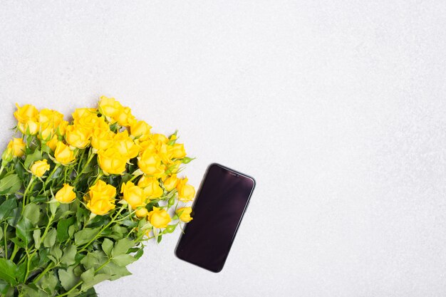 Frühlingssommerkonzept mit gelben Rosenblumen und Telefon auf weißem Tischhintergrund