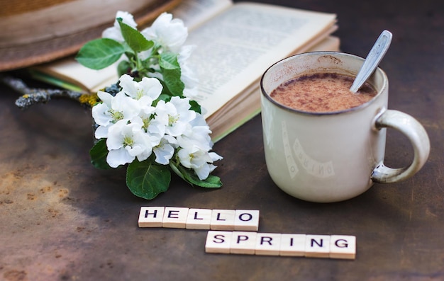 Frühlingsstillleben auf dunklem Hintergrund. Die Aufschrift "Hallo Frühling" Eine Tasse Kaffee, ein Buch, ein blühender Zweig eines Apfelbaums und ein Hut. Foto im Vintage-Stil, Blumenarrangement mit Zweig