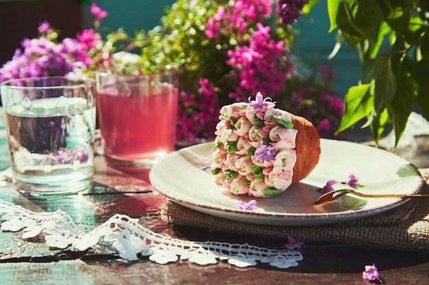 Frühlingstisch im Freien mit Blumenästhetik, Cupcake-Kompott und Glas Wasser Frühlingsfliederblumen im Freien an einem sonnigen Tag Schöpfer eines verträumten Dessertkonzepts