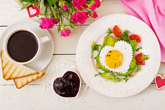 Frühstück am Valentinstag - Sandwich aus Spiegelei in Form eines Herzens, Avocado und frischem Gemüse. Tasse Kaffee. Englisches Frühstück. Draufsicht