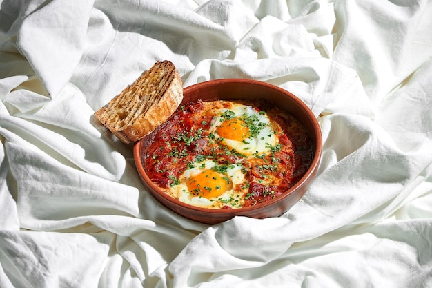 Foto frühstück im bett rührei mit shakshouka-tomaten in einer tonschale auf einem bettlaken