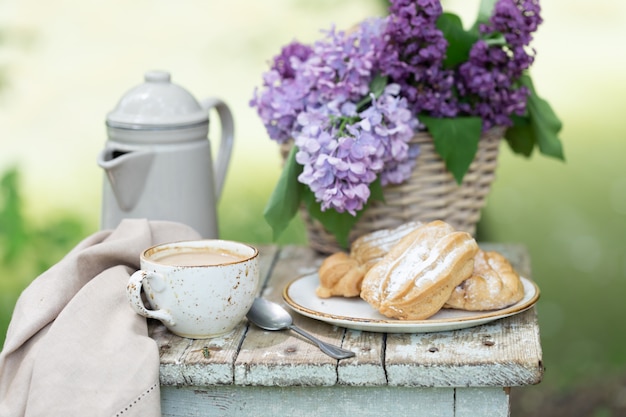 Frühstück im Garten: Eclairs, eine Tasse Kaffee, eine Kaffeekanne, lila Blumen in einem Korb.