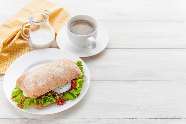 Frühstück im rustikalen Stil auf einer weißen Holzoberfläche. Ciabatta mit pochiertem Ei und Körnersenf mit einer heißen Tasse Kaffee und frisch