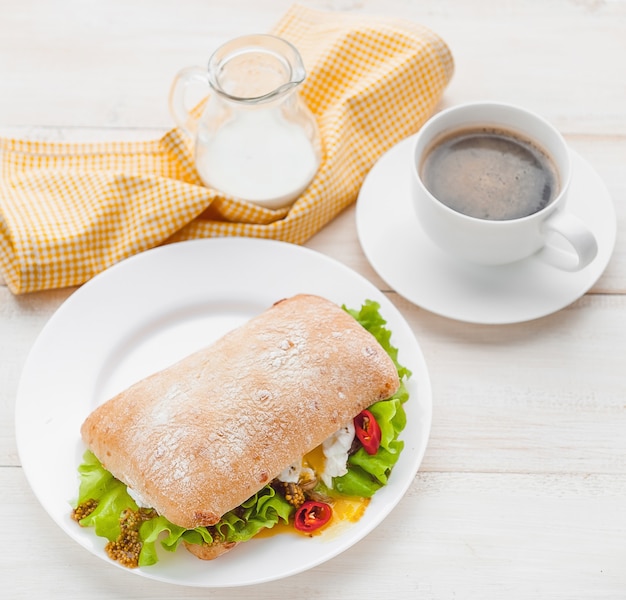 Frühstück im rustikalen Stil auf einer weißen Holzoberfläche. Ciabatta mit pochiertem Ei und Körnersenf mit einer heißen Tasse Kaffee und frisch