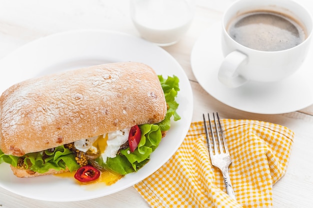 Frühstück im rustikalen Stil auf einer weißen Holzoberfläche. Ciabatta mit pochiertem Ei und Körnersenf mit einer heißen Tasse Kaffee und frisch