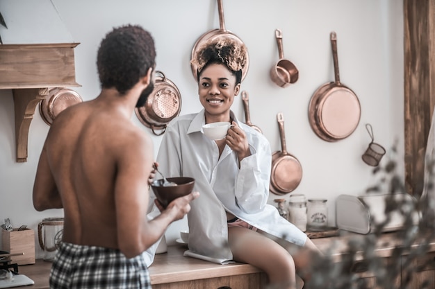 Frühstück, Kommunikation. Interessierte Afroamerikanerfrau im weißen Hemd mit Tasse Kaffee und Mann ohne Hemd