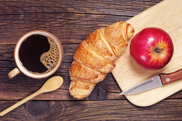 Frühstück mit Croissant, schwarzem Kaffee und Äpfeln auf einem dunklen Holztisch