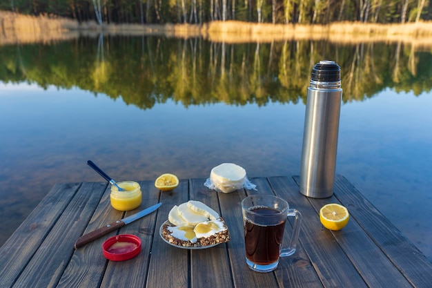 Frühstück mit heißem Tee, Käsesandwich und Honig auf Holztisch am Morgen neben dem See Natur- und Lebensmittelkonzept