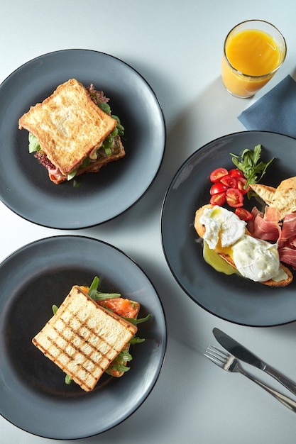 Frühstück mit Sandwiches auf Tellern, Benedict-Eiern mit Tomaten und Schinken mit Besteck und frischem Orangensaft