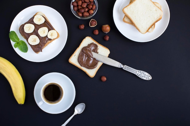 Frühstück mit Schokoladencremebrot, Banane und Kaffee auf schwarzem Hintergrund Draufsicht Kopierbereich