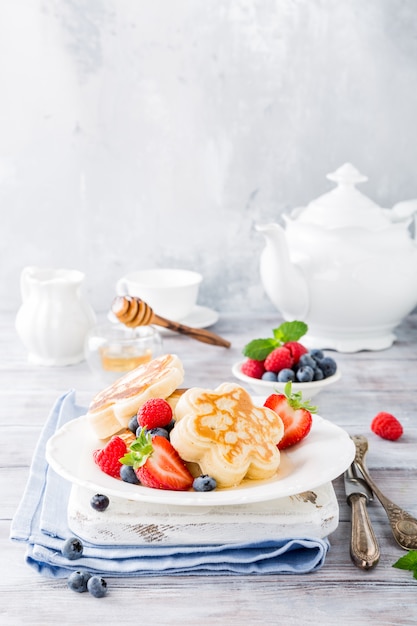 Frühstück mit schottischen Pfannkuchen in Blütenform, Beeren und Honig auf hellem Holztisch