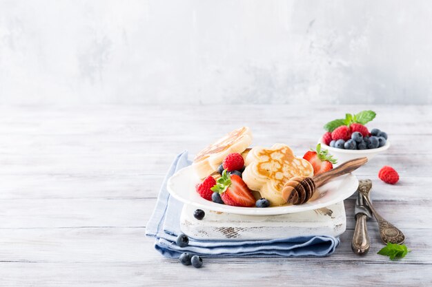 Frühstück mit schottischen Pfannkuchen in Blütenform, Beeren und Honig auf hellem Holztisch