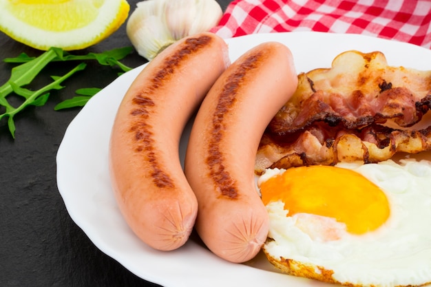 Frühstück mit Spiegeleiern, Speck, Würstchen und Gemüsesalat auf dunklem Steinhintergrund.