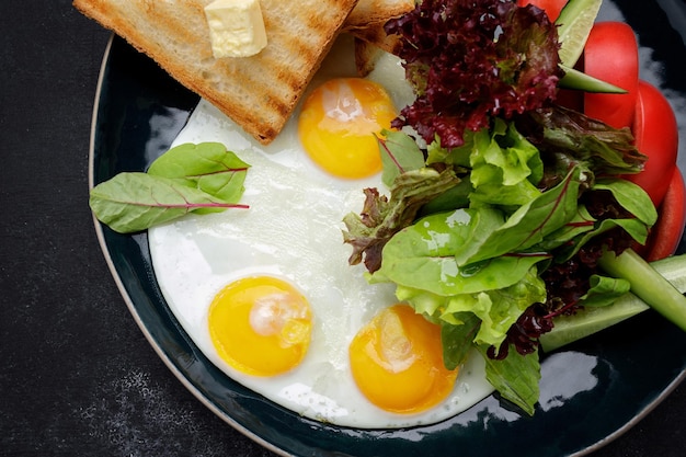 Frühstück Spiegeleier mit Toast mit Butter und Gemüse Gurken und Tomaten auf einem dunklen Teller vor einem dunklen Hintergrund