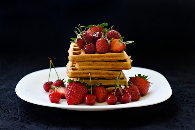 Frühstück. Waffeln mit Erdbeeren, Kirschen auf einer weißen Platte auf einem schwarzen Hintergrund