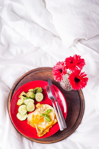 Frühstückseier auf einem weißen Teller mit halber Avocado, Gurken auf einem roten Teller auf dem Bett mit Blumen