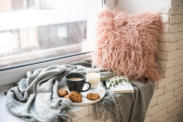Frühstückstasse im skandinavischen Stil mit Kaffee und Keksen auf gemütlicher Fensterbank mit warmer Decke