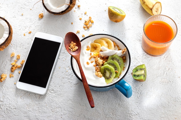 Frühstückstischkonzept. Smartphone mit Kokos-Joghurt mit Müsli und Früchten auf leichtem Tisch mit Karottensaft