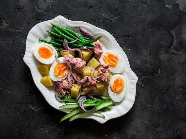 Frühstücksvorbereitungen Snack-Teller gekochte Eier Kartoffeln Stringbohnen rote Zwiebeln Gurken Konserven Thunfisch Salat auf einem dunklen Hintergrund Top-View