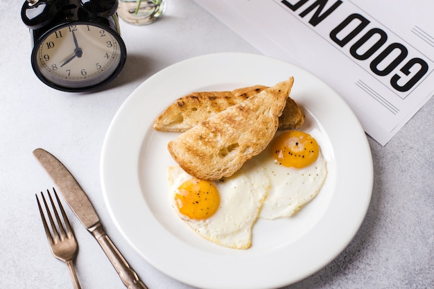 Frühstückszeit. Zwei Spiegeleier mit Toast zum Frühstück auf einem strukturierten hellgrauen Hintergrund mit einer Zeitung und einem Wecker