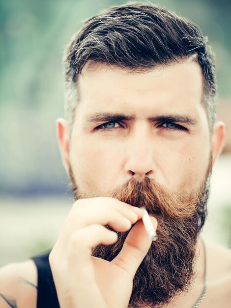 Fruncer el ceño apuesto hombre barbudo con bigote de barba y cabello gris con estilo hipster macho fumar cigarrillo al aire libre primer plano